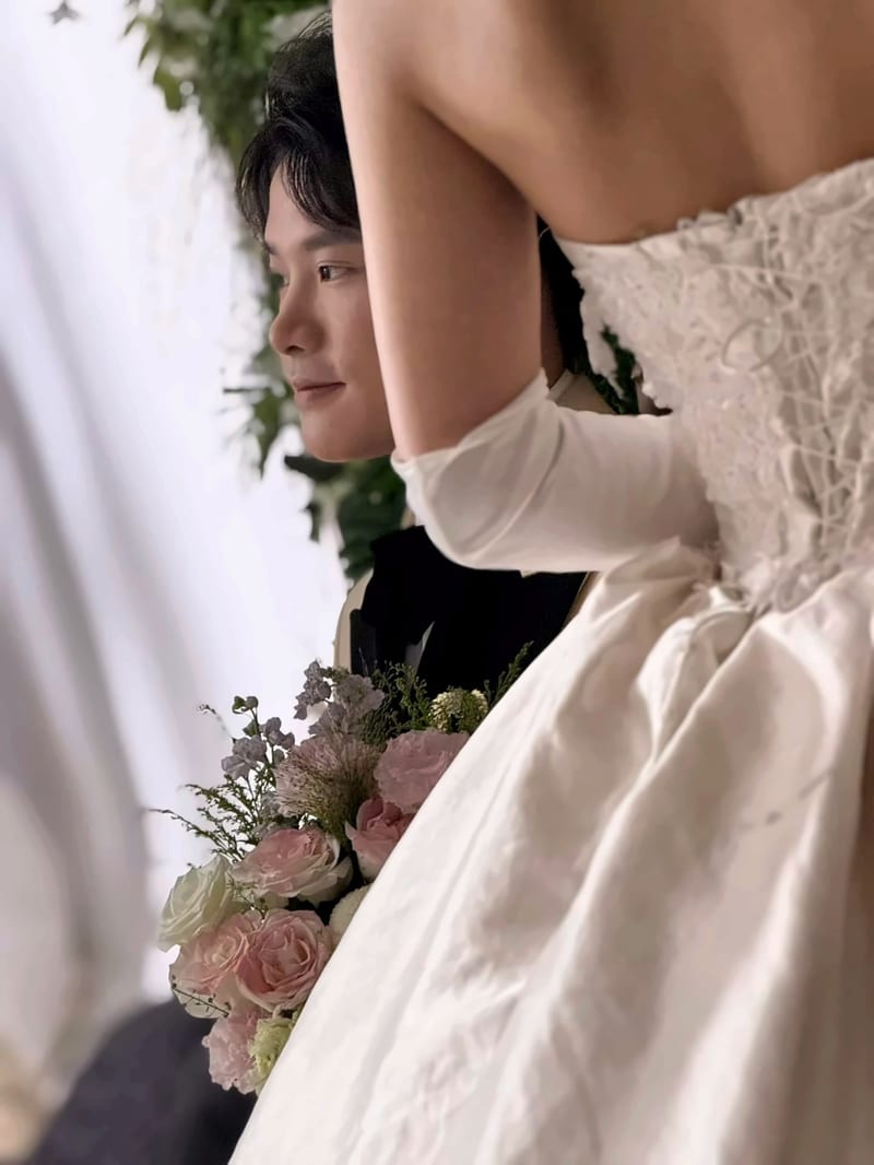 婚礼现场，白色蕾丝礼服与粉色花束占据前景，后方一名穿深色衣服的人侧身站立